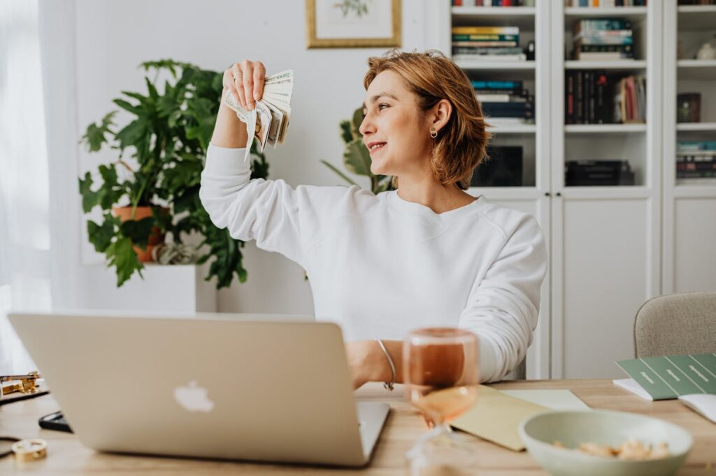 pexels photo 5899175 Smiling woman holding cash, working at home office with a laptop, symbolizing financial success.